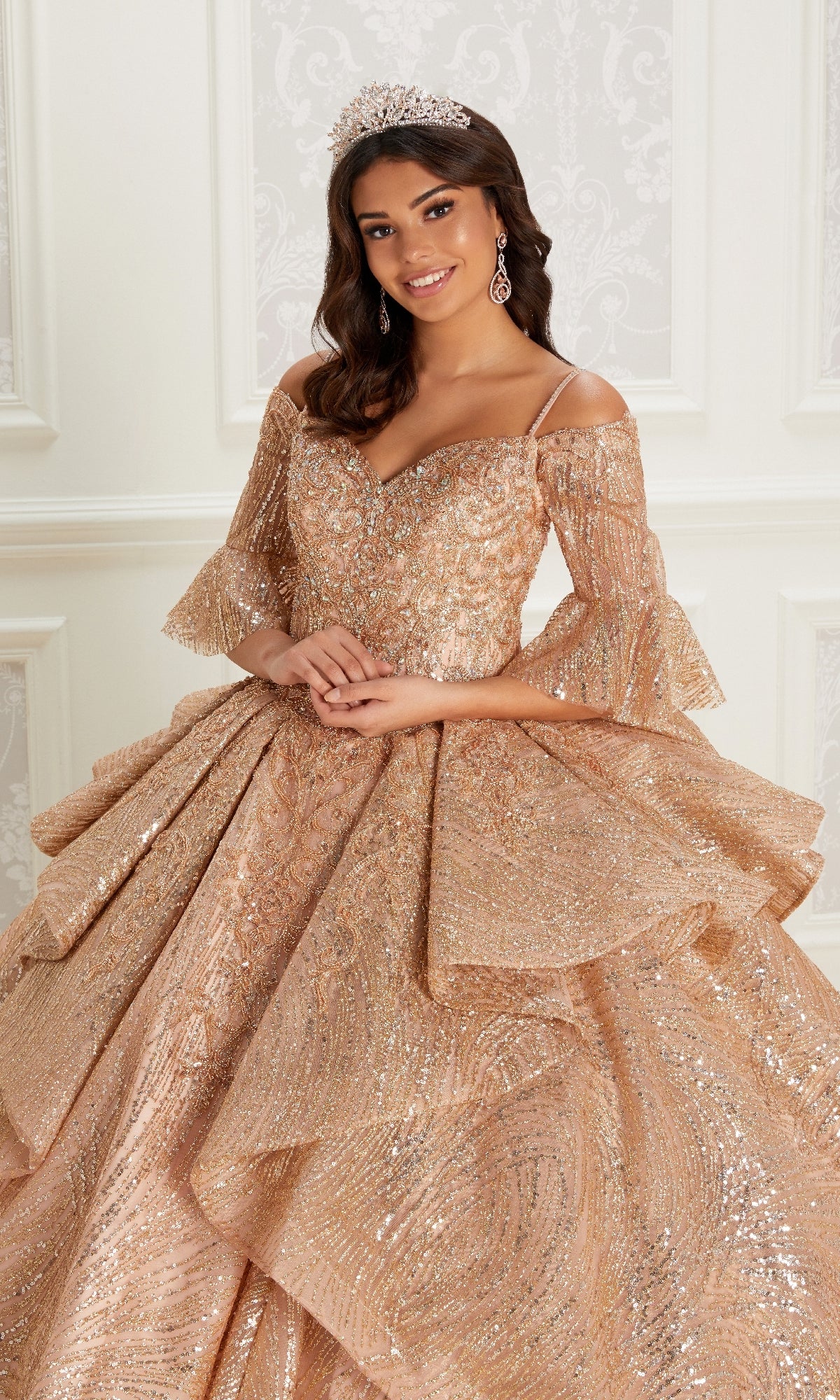 A woman stands indoors wearing the Quinceanera Dress PR22142 By Princesa, featuring sparkling gold fabric, layered ruffles, off-the-shoulder detachable sleeves, a corset back, and accessorized with a tiara and dangling earrings.
